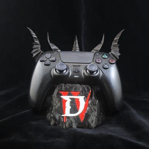 Puede incluir: Mando negro de PlayStation 5 personalizado con alas de murciélago y una espiga central. El mando descansa sobre una base de piedra con el logotipo "D IV" en blanco y rojo. Fondo oscuro.