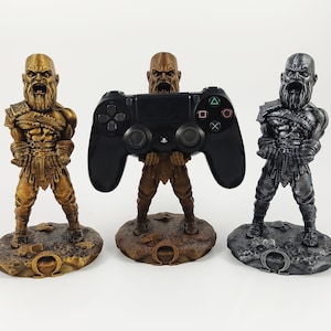 May include: Three Kratos figures holding a black PlayStation controller. The figures are made of plastic and are painted in gold, bronze, and silver. Each figure stands on a base with the Greek letter Omega.