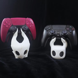 May include: Two video game controllers, one burgundy and one black, sit on white, stylized character stands. The burgundy controller is a PlayStation 5 DualSense, and the black controller is a PlayStation 4 DualShock 4.