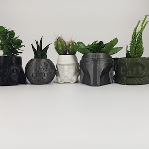 May include: Five small potted plants in black, white and green Star Wars character themed planters. The planters are shaped like Darth Vader, a Death Star, a Stormtrooper, Boba Fett and Baby Yoda.