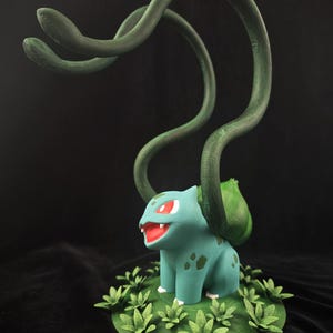 May include: A 3D printed figurine of a green and blue Pokemon character with red eyes and a green vine-like appendage on its back. The figurine is standing on a green base with green leaves.