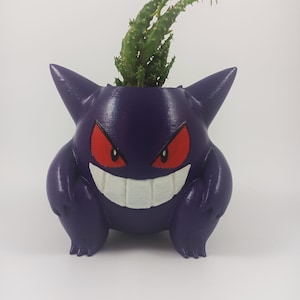 Puede incluir: Maceta Gengar de color púrpura impresa en 3D con una planta suculenta verde que crece en la parte superior.