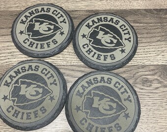 Könnte beinhalten: Vier runde Untersetzer mit den Worten "Kansas City Chiefs" und dem Teamlogo. Die Untersetzer sind grau mit einer strukturierten Oberfläche und einem dunkelgrauen Rand. Das Teamlogo ist ein roter Pfeil mit den Buchstaben "KC" darin.