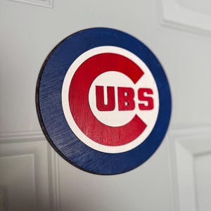 May include: A round wooden sign featuring the Chicago Cubs logo. The sign has a blue outer ring, a red inner circle, and a white border. The word "CUBS" is in red letters in the center.