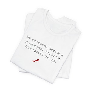 May include: White t-shirt featuring the quote "By all means, move at a glacial pace. You know how that thrills me." and a red high heel illustration. The shirt is folded, likely made of cotton.