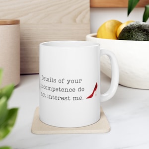 May include: White ceramic mug with a black and white text that reads "Details of your incompetence do not interest me." A red high heel shoe is pictured next to the text.