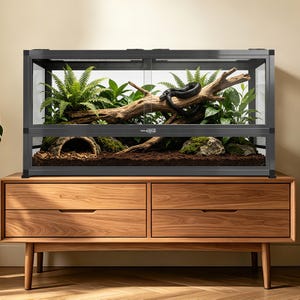 May include: A large, rectangular reptile enclosure with a black frame and sliding glass doors. Inside, a black snake rests on a large piece of driftwood surrounded by lush green plants. The enclosure sits atop a wooden cabinet with four drawers.