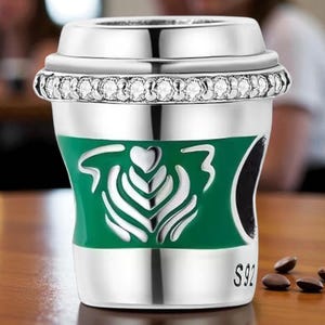 Könnte beinhalten: Silberfarbener Kaffeebecher-Charm mit einem grünen Band mit einem weißen Herz- und Blattdesign. Der Rand ist mit klaren Kristallen verziert. Der Charm befindet sich neben Kaffeebohnen auf einer Holzoberfläche.