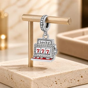 May include: A silver charm shaped like a slot machine with the word "Lucky" above the reels. The reels display the number "777" in red. The charm is embellished with clear crystals and hangs from a decorative ring.