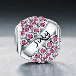May include: A silver charm with the word "July" in cursive. The charm is adorned with pink gemstones in a repeating pattern. The charm is a bead, likely for a bracelet or necklace.
