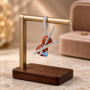 Puede incluir: Un dije de plata con forma de violín, cuerpo marrón rojizo, arco de plata y una pequeña gema azul en forma de corazón en la base. El dije cuenta con una argolla de plata incrustada con piedras transparentes, colgado de un soporte dorado.