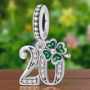 May include: Silver charm with the number 20, adorned with clear crystals and a green four-leaf clover. The charm has a loop with heart-shaped cutouts for attaching to a bracelet or necklace. The background is a blurred view of greenery and flowers.