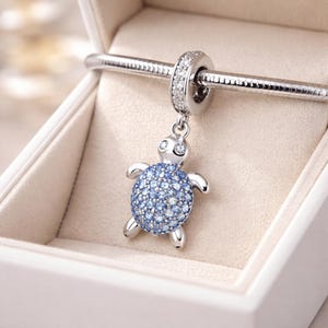 May include: A silver turtle charm with a shell of blue stones. The charm hangs from a silver ring studded with clear stones. The turtle charm is presented in a white jewellery box. The charm is suitable for a bracelet.