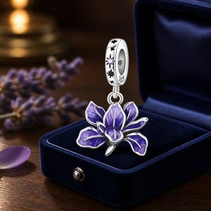Pode incluir: Um berloque de prata com um pendente de flor esmaltada em roxo suspenso por uma argola cilíndrica. A argola é decorada com recortes de estrelas pretas e uma pequena pedra roxa, com a marca S925 visível na lateral do metal.