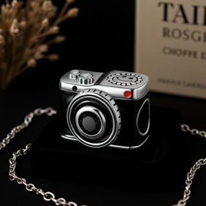 May include: A black and silver camera-shaped lighter with a smiley face design on top. The lighter has a lens-like feature and a red button. A silver chain is in the foreground.