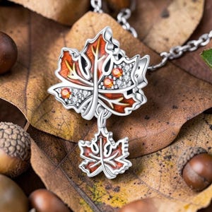 Può includere: Collana in argento con ciondolo a forma di foglia d'acero. Il ciondolo presenta un design dettagliato con accenti rossi, arancioni e bianchi. La collana è ambientata su uno sfondo di foglie autunnali e ghiande.