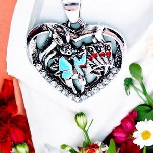 May include: A silver heart-shaped pendant necklace with a detailed design of a rabbit holding playing cards. The rabbit is wearing a blue vest and the cards are red and black.