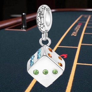 May include: A silver charm with a dice design. The dice has colorful gems and is suspended from a loop with star accents. The charm is set against a casino table background.