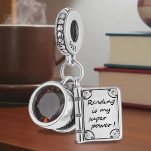 Op de afbeelding: Een zilveren bedel met een koffiekopje en een boek met de tekst "Reading is my super power!". De beker heeft een donkerbruine edelsteen. De bedel hangt voor een bruin koffiekopje en boeken.