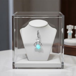 Puede incluir: Un charm plateado con forma de luciérnaga, con un centro azul brillante, exhibido sobre un busto blanco dentro de una caja de acrílico transparente. El charm de la luciérnaga tiene una corona y detalles de diamantes. El charm es un colgante.