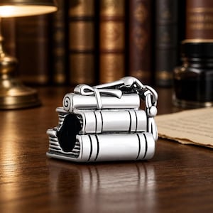 May include: A silver charm shaped like a stack of three books with a rolled scroll on top. The charm has a detailed design with lines representing pages and a clasp. The background features a desk and a collection of books.