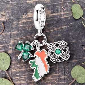 May include: A silver charm featuring a cylindrical bead engraved with St. Patrick's Day S925. Three dangling pendants are attached: a three-leaf clover with green stones, a map of Ireland in orange, white, and green enamel, and a Celtic cross.