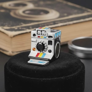 May include: A silver-colored camera ring with a black lens and colorful accents. The ring features a detailed design with a black velvet base. The camera has a black gem and two clear gems.