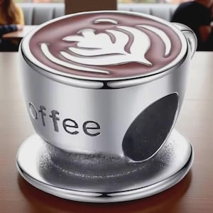 Puede incluir: Un charm plateado con forma de taza de café con la palabra "coffee" grabada en el lateral. La parte superior de la taza presenta un diseño de arte latte marrón y blanco. El charm se apoya en un platillo a juego.