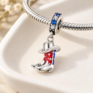 Könnte beinhalten: Ein silberner Charm-Anhänger in Form eines Cowboystiefels mit rot-weiß-blauer Emaille-Verzierung und kleinen Sternausschnitten. Der Anhänger ist auf einer silbernen Schlangenkette befestigt und mit abwechselnd blauen und klaren Steinen besetzt.