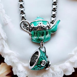 Könnte beinhalten: Halskette mit einem silberfarbenen Anhänger in Form einer türkisfarbenen Teekanne und einem passenden Tassen-Charm. Die Teekanne und die Tasse haben silberne Verzierungen und sind an einer Kette befestigt.