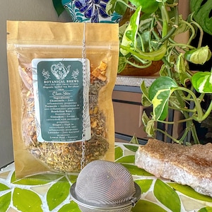 May include: A clear plastic bag of herbal tea labeled "Botanical Brews Clear Skin" with a metal tea infuser. The tea bag contains dried herbs and spices. The tea infuser is round and made of a fine mesh. The background includes green plants and a decorative tile surface.