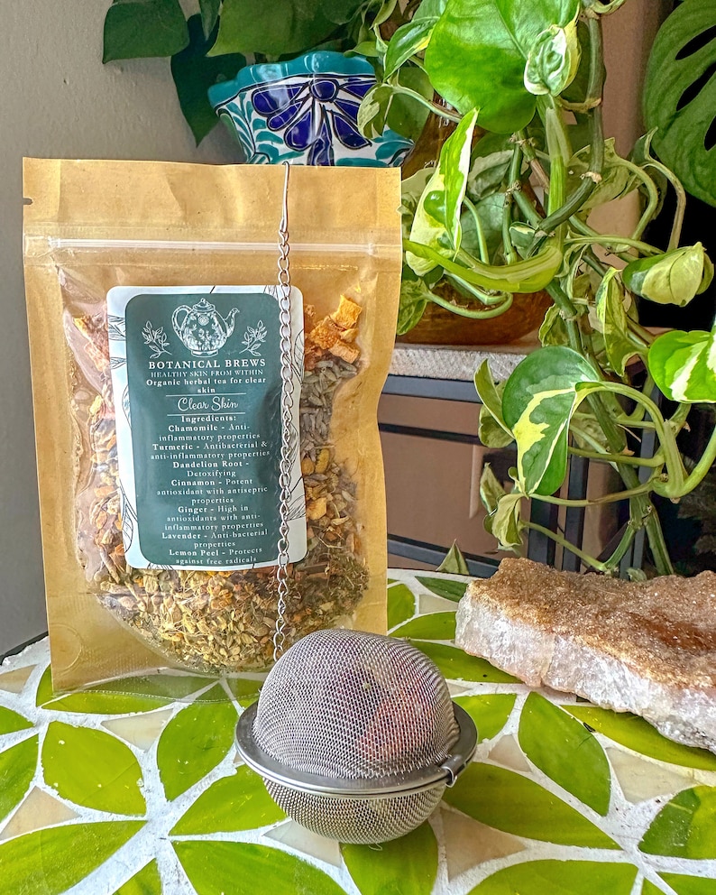 May include: A clear plastic bag of herbal tea labeled "Botanical Brews Clear Skin" with a metal tea infuser. The tea bag contains dried herbs and spices. The tea infuser is round and made of a fine mesh. The background includes green plants and a decorative tile surface.