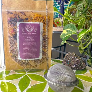 May include: A clear bag of herbal tea with a purple label that reads "BOTANICAL BREWS". A stainless steel tea infuser sits in front of the bag. The tea infuser is round and has a chain. The background includes plants and a decorative tile surface.