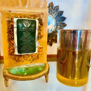 May include: A clear glass tea infuser and a bag of herbal tea labeled "Botanical Brews" for clear skin. The bag is filled with dried herbs and spices and sits on a gold-colored stand. The tea infuser is made of glass and metal.