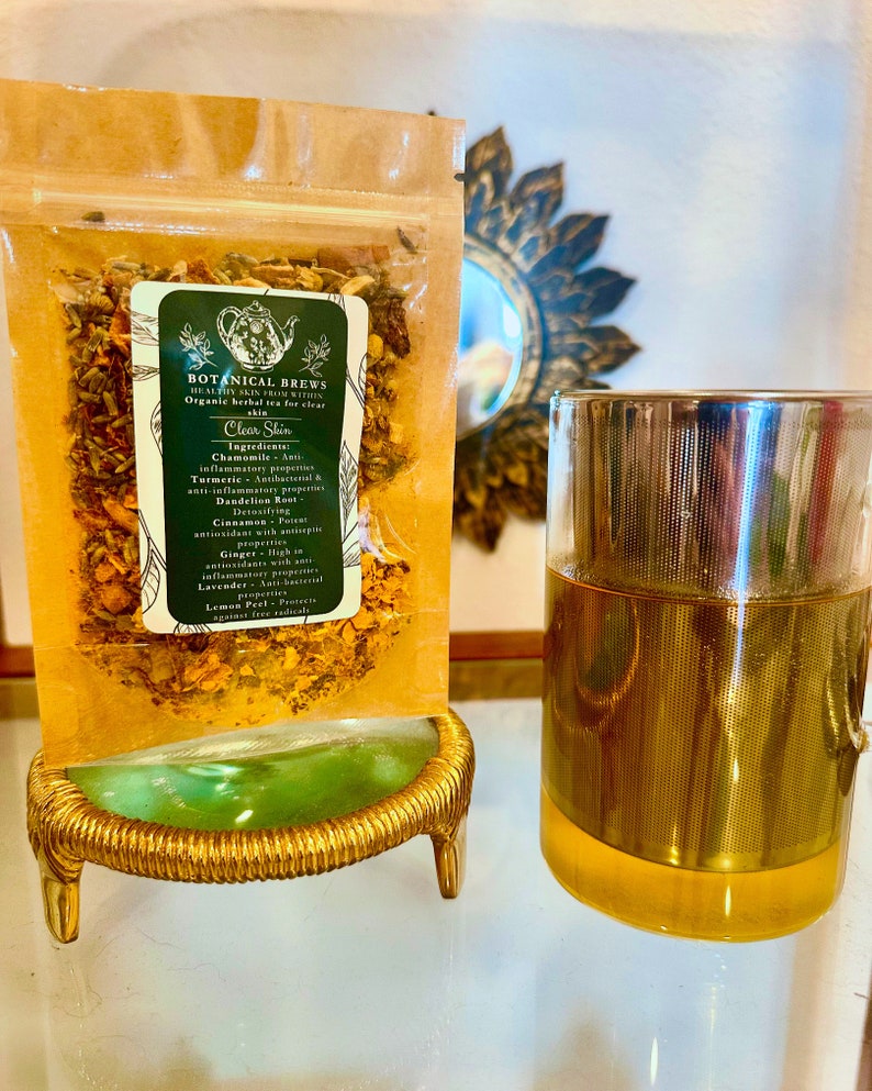 May include: A clear glass tea infuser and a bag of herbal tea labeled "Botanical Brews" for clear skin. The bag is filled with dried herbs and spices and sits on a gold-colored stand. The tea infuser is made of glass and metal.