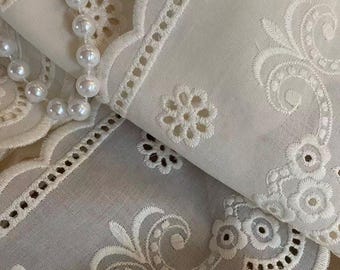 May include: Off-white cotton fabric featuring intricate eyelet embroidery with floral patterns and scalloped edges. A strand of white faux pearls is draped across the folded textile, highlighting the detailed needlework and cut-out designs.
