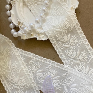May include: White lace ribbon with a floral rose pattern. A string of white pearls and a decorative white plate are in the background. A clear iridescent butterfly is in the foreground.