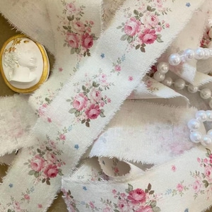 May include: A length of off-white fabric ribbon with frayed edges, featuring a repeating pattern of pink roses and small green leaves. A white oval cameo brooch with a gold rim and a strand of white faux pearls are positioned near the ribbon.