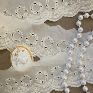 May include: White eyelet lace trim with a scalloped edge and floral cutouts. A white cameo with a gold frame and a strand of white pearls are also in the image. The lace is a delicate fabric, perfect for sewing and crafting projects.