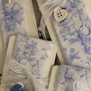 May include: Several strips of white fabric featuring a blue printed pattern of rabbits, mushrooms, and flowers. Each strip is wrapped around a card and decorated with white lace ribbon, small blue fabric bows, and heart-shaped buttons in blue or white.