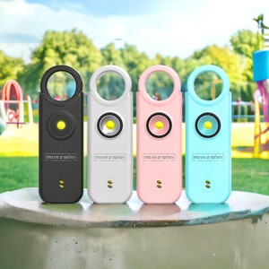 May include: Four personal safety alarms in black, white, pink, and light blue, each with a circular top loop and a central speaker. The alarms have a yellow light and the text "PRIAM & BJÖRN ESTABLISHED 2011".