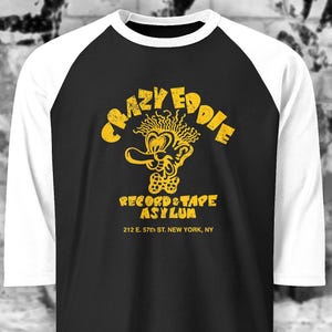 Op de afbeelding: Zwart en wit raglan baseball t-shirt met een gele afbeelding. De afbeelding leest "Crazy Eddie Record & Tape Asylum" met een stripfiguur en het adres "212 E. 57th St. New York, NY."