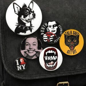 May include: A black canvas messenger bag decorated with six circular pin-back buttons. The buttons feature a black and white chihuahua, a person holding a red mug with text, a smiling person, a red mouth with fangs and text, and a yellow cat face.