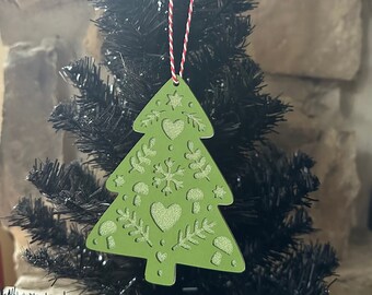 May include: A flat, olive green, tree-shaped ornament featuring cut-out designs of hearts, snowflakes, mushrooms, and sprigs. The cut-out areas have a shimmering, glittery texture. It hangs from a red and white twisted string loop.