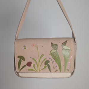 May include: A light pink shoulder bag with a cream-colored bottom and strap. The bag features a botanical illustration of carnivorous plants in green, pink, and purple. The bag has a rectangular shape.