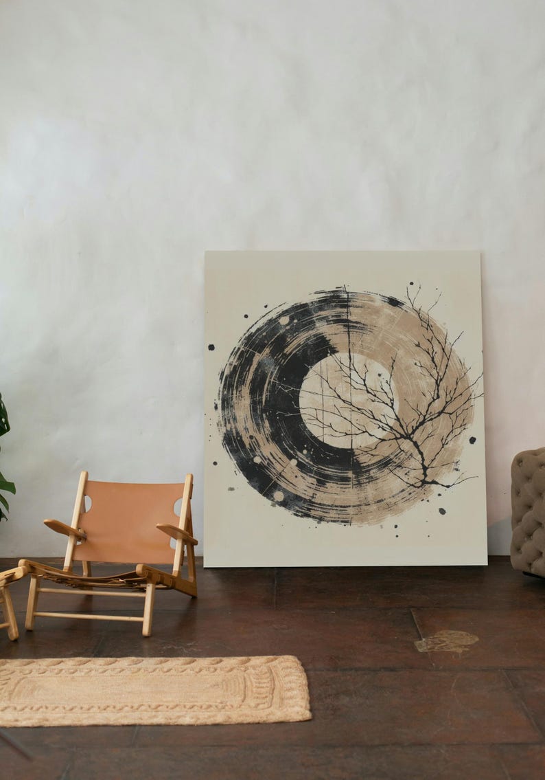 Op de afbeelding: Abstract kunstcanvas met een zwart en beige cirkelvormig ontwerp en een illustratie van een boomtak. Een lichtbruine leren stoel en een geweven vloerkleed staan op de voorgrond. Het kunstwerk wordt tentoongesteld in een kamer met een neutraal kleurenpalet.