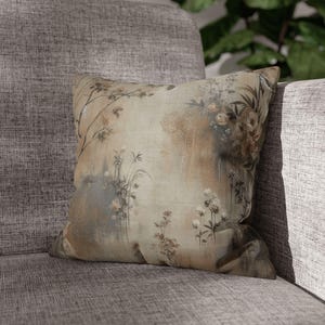 May include: Square decorative pillow with a floral design in muted tones of beige, brown, and gray. The pillow features a botanical print with flowers and foliage, resting on a gray upholstered chair.