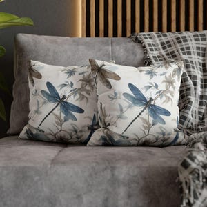 May include: Two decorative throw pillows with a dragonfly design. The pillows feature a white background with blue and brown dragonflies and foliage. The pillows are placed on a gray sofa, with a plaid blanket in the background.
