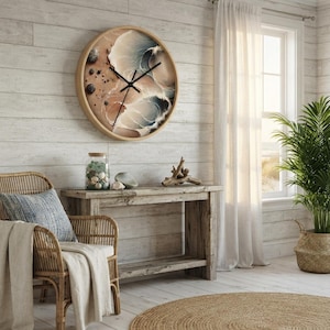 May include: A round wall clock with a light wood frame featuring an abstract resin art face in shades of beige, cream, and dark blue, with black clock hands. The design includes small dark pebbles on the left side of the clock face.