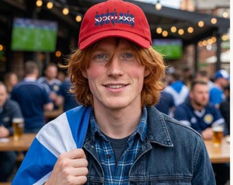 May include: A red baseball cap featuring the word SCOTLAND printed in a stylised blue and white font across the front panel. The cap has a curved brim and a structured crown, designed with a classic six-panel construction and a button on top.